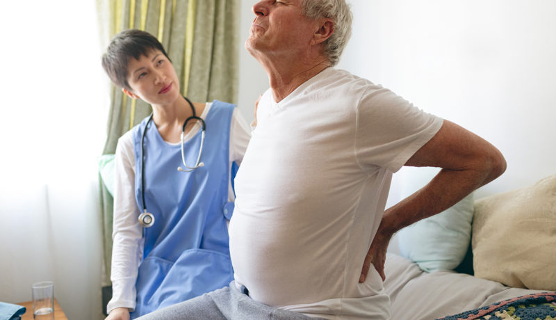 Side view of Asian female nurse helping senior Caucasian male patient for back pain at retirement home