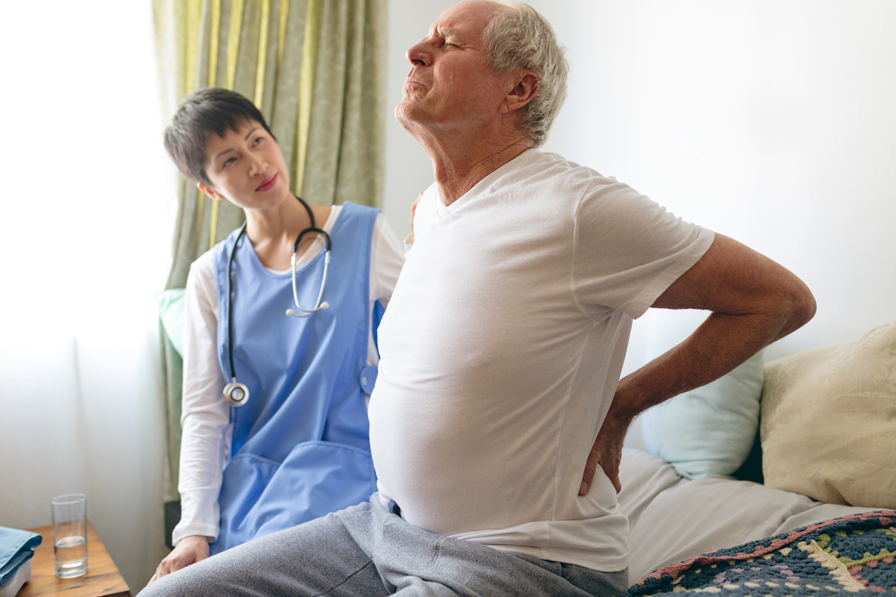 Side view of Asian female nurse helping senior Caucasian male patient for back pain at retirement home